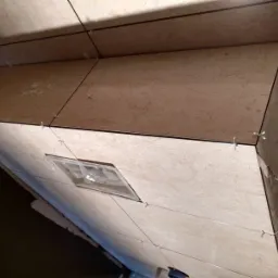 Świeżo ułożone, beżowe płytki z delikatnym wzorem marmuru na zabudowie, z widocznymi krzyżykami dystansowymi i czarną fugą, częściowo oświetlone przez otwór rewizyjny.