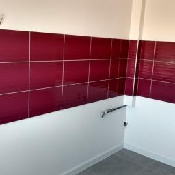 Prestige Home - Ściana w trakcie wykańczania z bordowymi, błyszczącymi płytkami ceramicznymi ułożonymi do połowy wysokości, widoczne puszki elektryczne i rury instalacyjne, biała farba na ścianie i szare płytki...