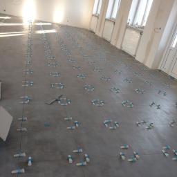 Prestige Home - Szare płytki podłogowe w trakcie układania, z użyciem niebieskich klinów poziomujących, w jasnym pomieszczeniu z oknami.