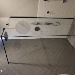 Nowoczesna kabina prysznicowa z czarnymi elementami, szklaną ścianą i szarymi płytkami ułożonymi w jodełkę na podłodze i ścianach, widoczne elementy instalacji wodnej.