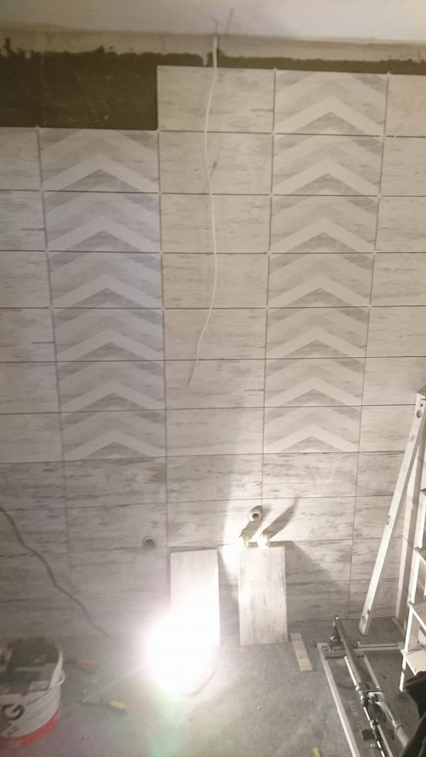 Ściana w trakcie układania płytek z motywem jodełki, widoczne niedokończone fragmenty i narzędzia pracy, w tle drabina i wiadro z zaprawą.