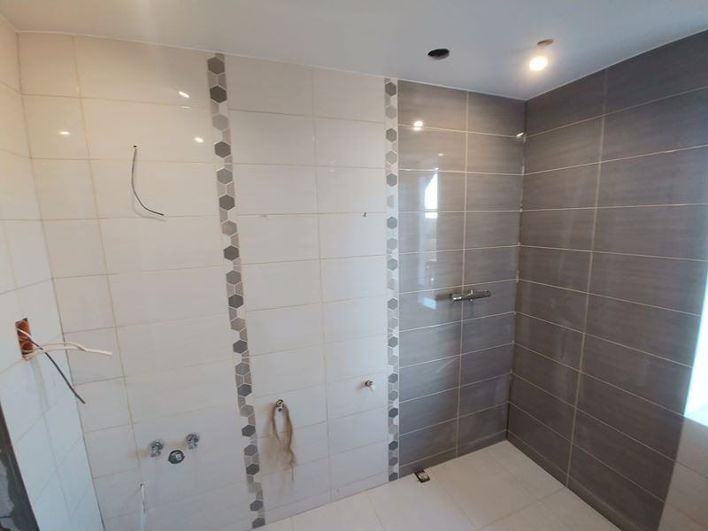 Wykończenie łazienki: Szare i białe płytki ceramiczne ułożone na ścianach, z dekoracyjnym pasem z heksagonów. Widoczne niedokończone podłączenia elektryczne i hydrauliczne.