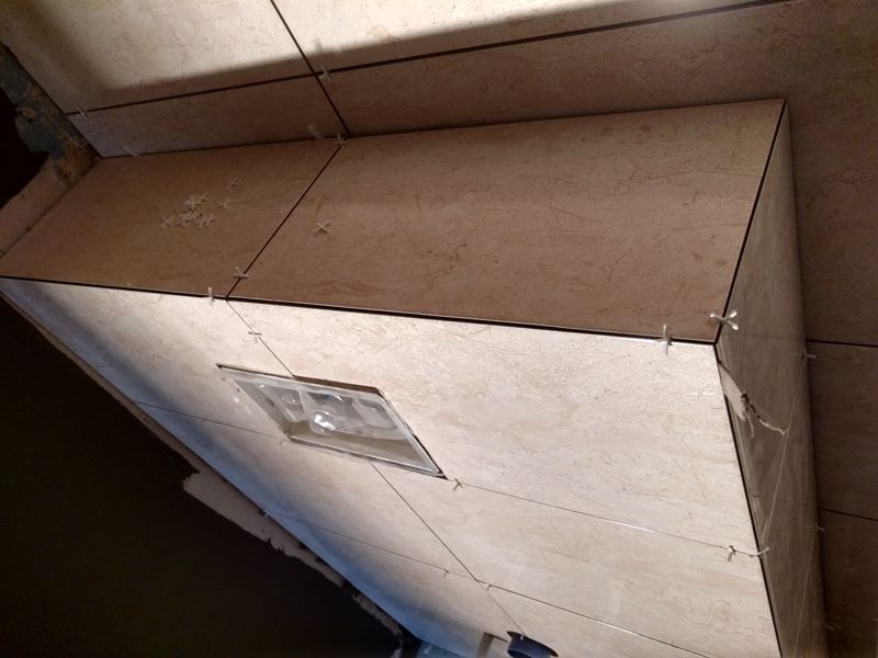 Świeżo ułożone, beżowe płytki z delikatnym wzorem marmuru na zabudowie, z widocznymi krzyżykami dystansowymi i czarną fugą, częściowo oświetlone przez otwór rewizyjny.