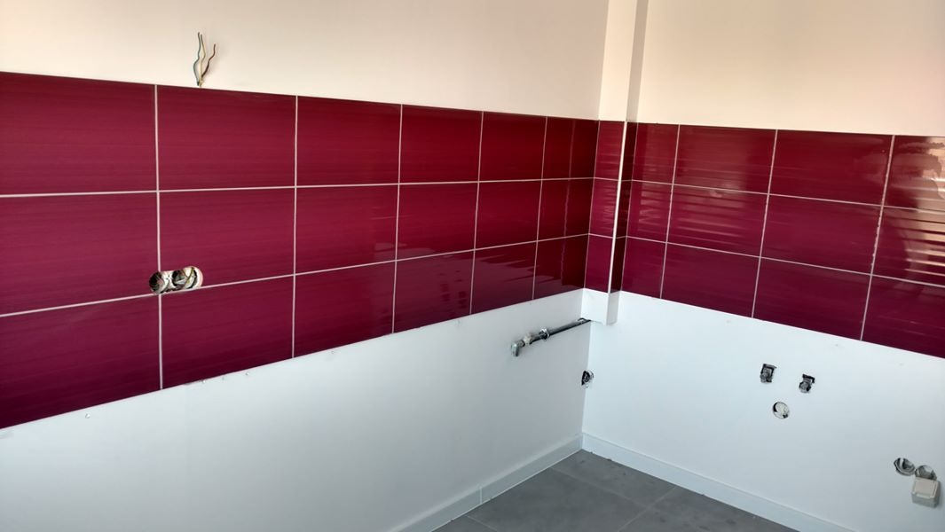 Ściana w trakcie wykańczania z bordowymi, błyszczącymi płytkami ceramicznymi ułożonymi do połowy wysokości, widoczne puszki elektryczne i rury instalacyjne, biała farba na ścianie i szare płytki...