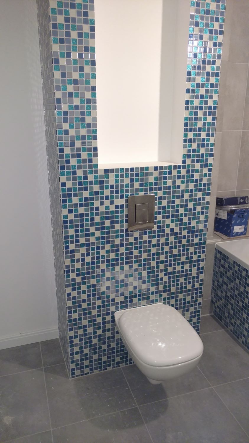 Wanna z obudową i toaleta podwieszana, wykończone mozaiką w odcieniach błękitu i szarości; widoczny fragment ściany z płytkami w neutralnym odcieniu szarości.