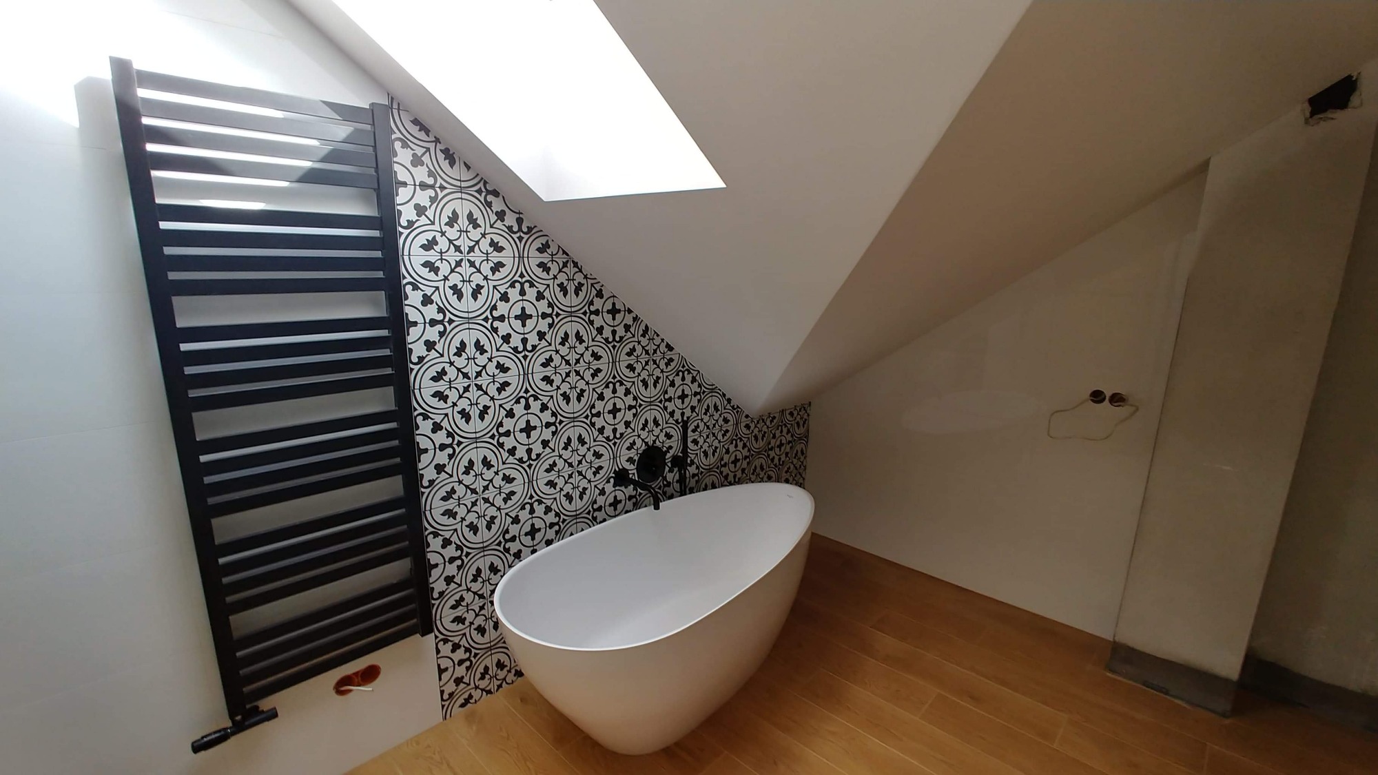 Biała, wolnostojąca wanna owalna z czarną armaturą na tle ściany wyłożonej czarno-białymi płytkami wzorzystymi w łazience na poddaszu z drewnianą podłogą i grzejnikiem drabinkowym.