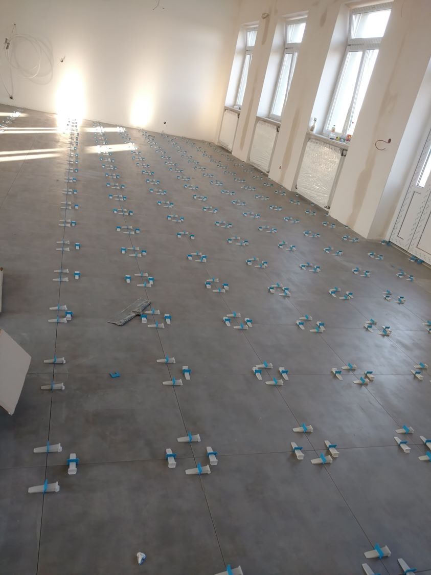 Szare płytki podłogowe w trakcie układania, z użyciem niebieskich klinów poziomujących, w jasnym pomieszczeniu z oknami.