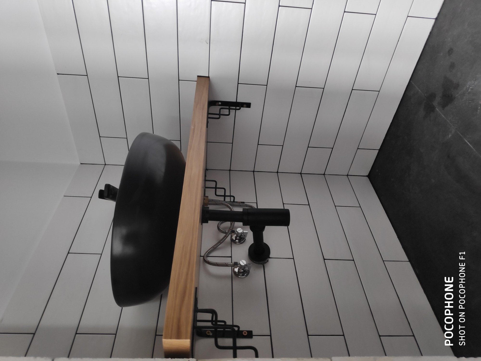 Czarna, okrągła umywalka nablatowa zamontowana na drewnianym blacie z czarnymi wspornikami, widoczne białe płytki ułożone w cegiełkę i czarna ściana w tle.