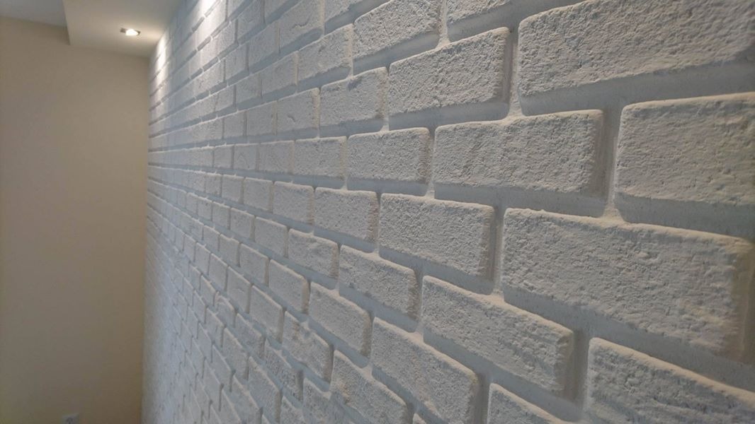 Ściana wykończona dekoracyjną, białą cegłą, z widoczną fakturą i fugami, oświetlona punktowym światłem z góry.