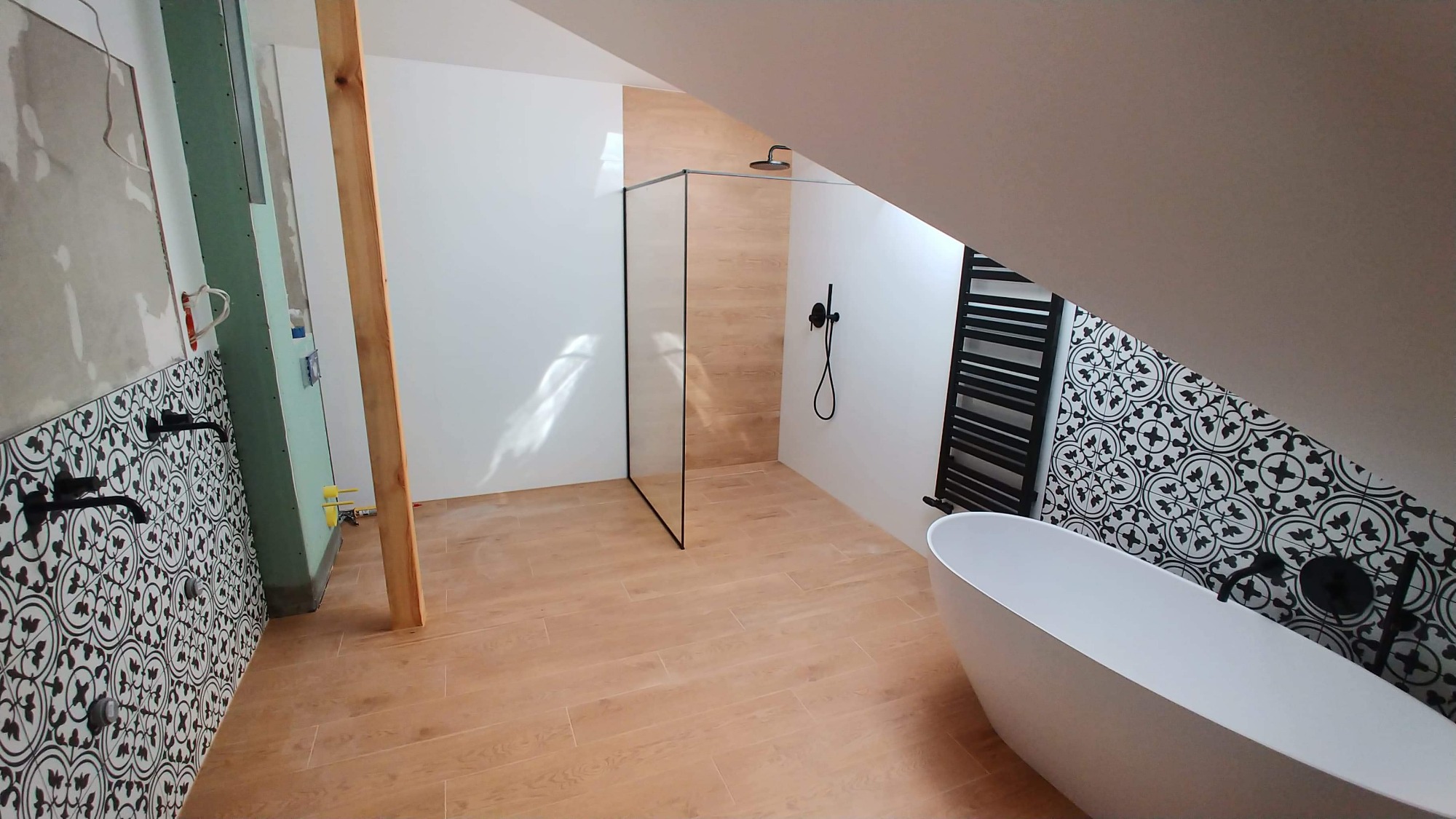 Wnętrze łazienki na poddaszu w trakcie wykańczania: biała wolnostojąca wanna, przeszklona kabina prysznicowa, dekoracyjne czarno-białe płytki na ścianie, drewnopodobna podłoga, grzejnik drabinkowy,...