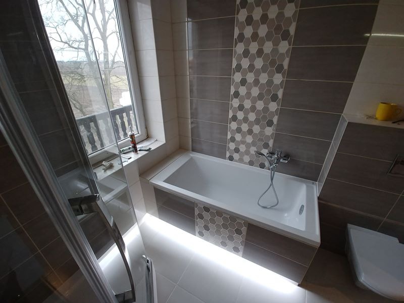 Nowoczesna łazienka z wanną podświetloną LED, szarymi płytkami z heksagonalnym wzorem i oknem z widokiem na drzewa; silikon sanitarny i narzędzia na parapecie.