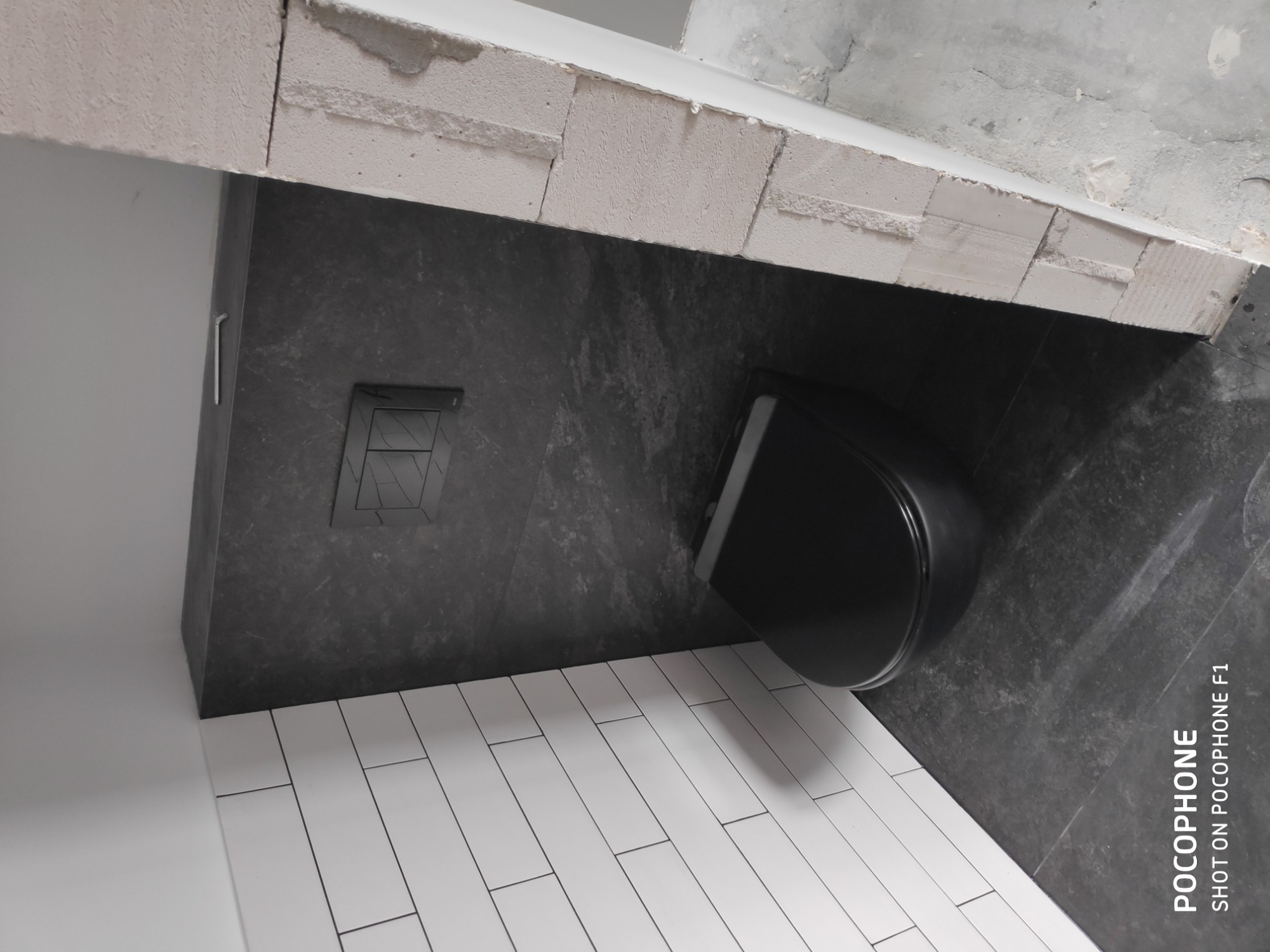 Częściowo wykończona łazienka z czarną miską WC, szarą ścianą z kamienia i białą podłogą z płytek w kształcie cegiełek, widoczne surowe wykończenie ściany nad miską WC.