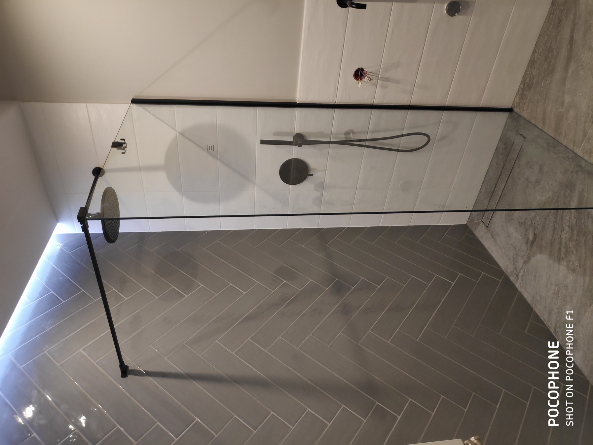 Nowoczesna kabina prysznicowa z czarnymi elementami, szklaną ścianą i szarymi płytkami ułożonymi w jodełkę na podłodze i ścianach, widoczne elementy instalacji wodnej.