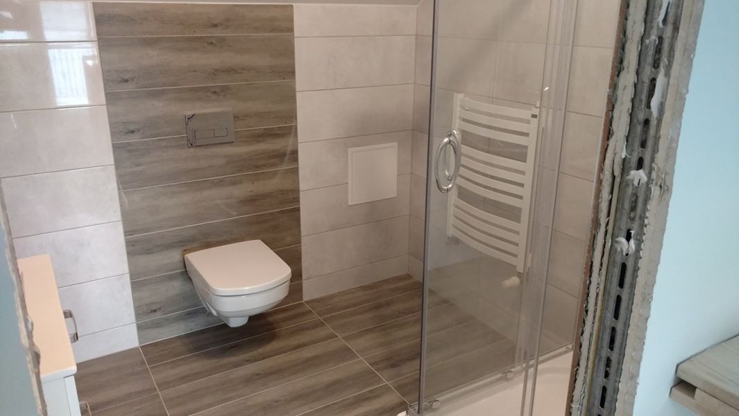 Nowoczesna łazienka z płytkami imitującymi drewno na podłodze i ścianie, podwieszana toaleta z szarą armaturą, biały grzejnik drabinkowy za szklaną kabiną prysznicową, widoczne surowe wykończenie...