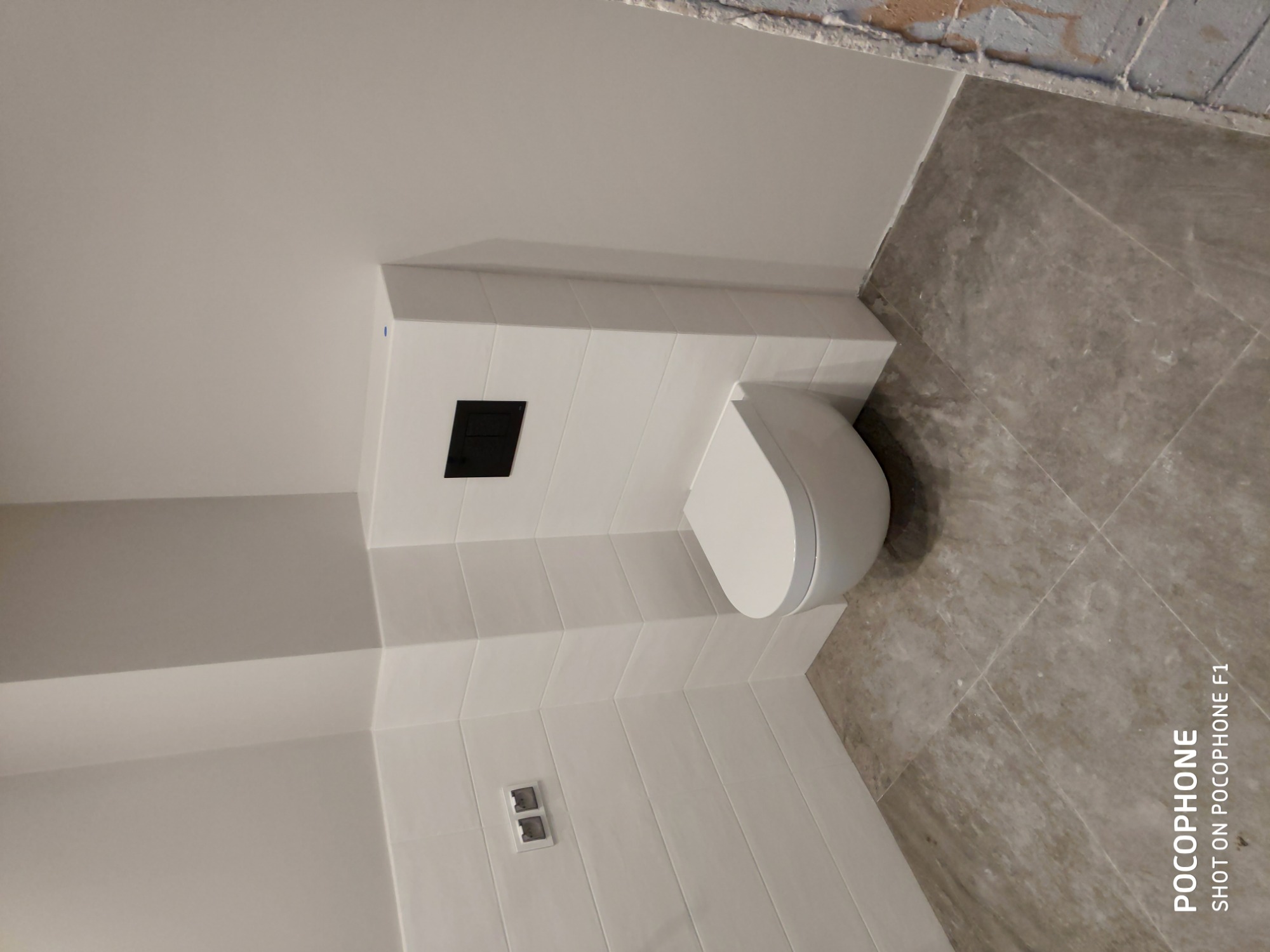 Nowoczesna toaleta podwieszana z czarnym przyciskiem spłukującym, obudowana białymi płytkami, szare płytki podłogowe, widoczne gniazdko elektryczne, surowy stan wykończenia sufitu.