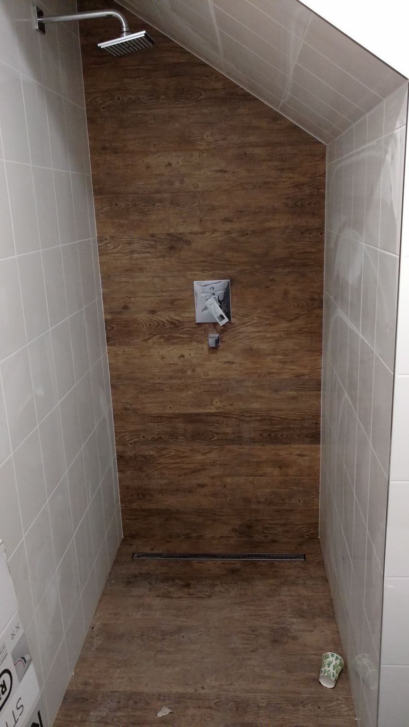 Kabina prysznicowa wykończona płytkami imitującymi drewno na ścianie i podłodze, z odpływem liniowym i baterią natryskową w kształcie kwadratu, białe płytki na bocznych ścianach, nachylony sufit.