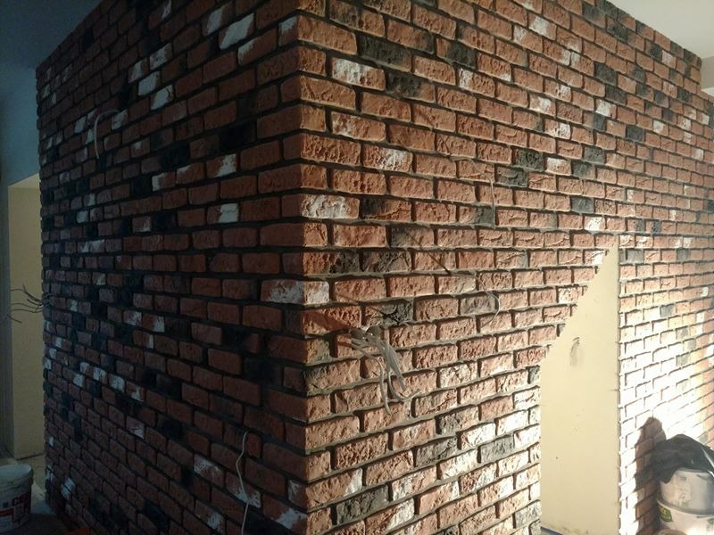 Ściana wykończona czerwoną cegłą klinkierową z nieregularnymi, ciemnymi wstawkami, widoczne przewody elektryczne i wiadro z farbą w dolnym rogu.