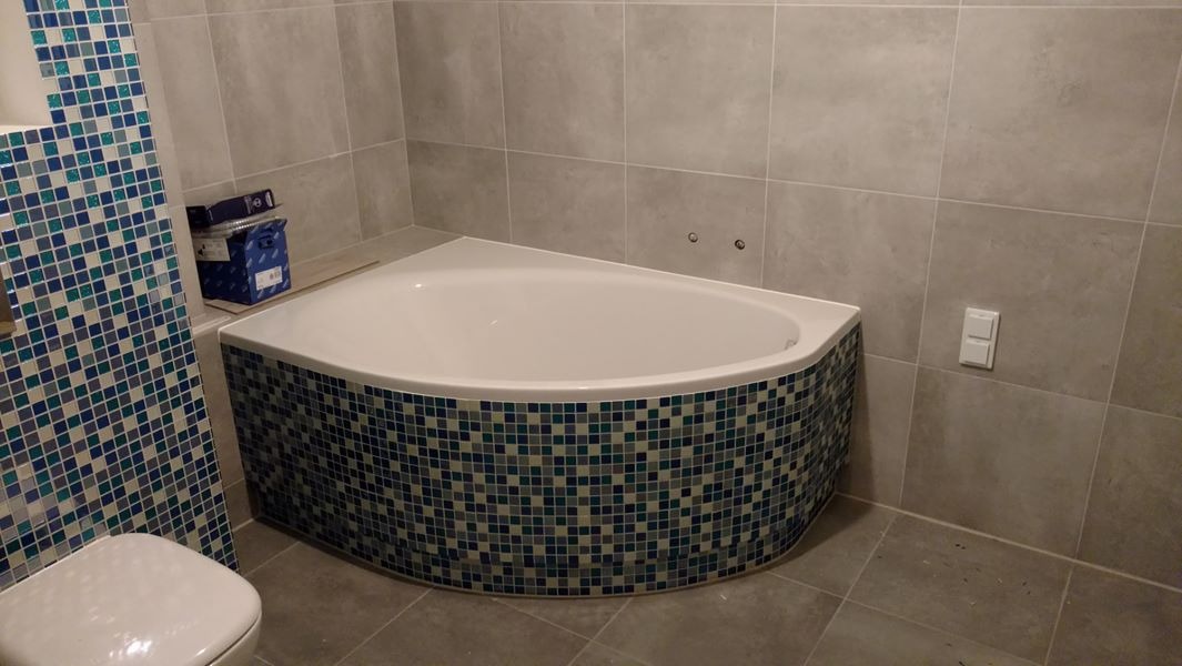 Narożna wanna obudowana mozaiką w odcieniach błękitu i szarości, dopasowana do mozaiki na ścianie obok toalety w wykończonej łazience z szarymi płytkami.