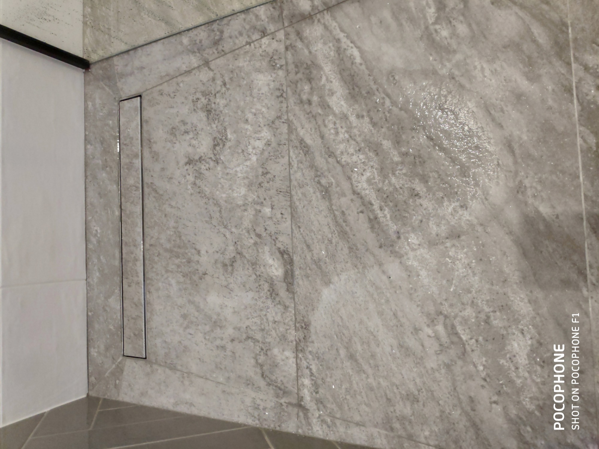 Szare, marmurkowe płytki w kabinie prysznicowej z liniowym odpływem, widoczne ślady wody na powierzchni płytek.