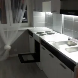 Jasna kuchnia z białymi szafkami, szarymi płytkami i otwartym piekarnikiem, oświetlona lampą wiszącą i światłem spod szafek.