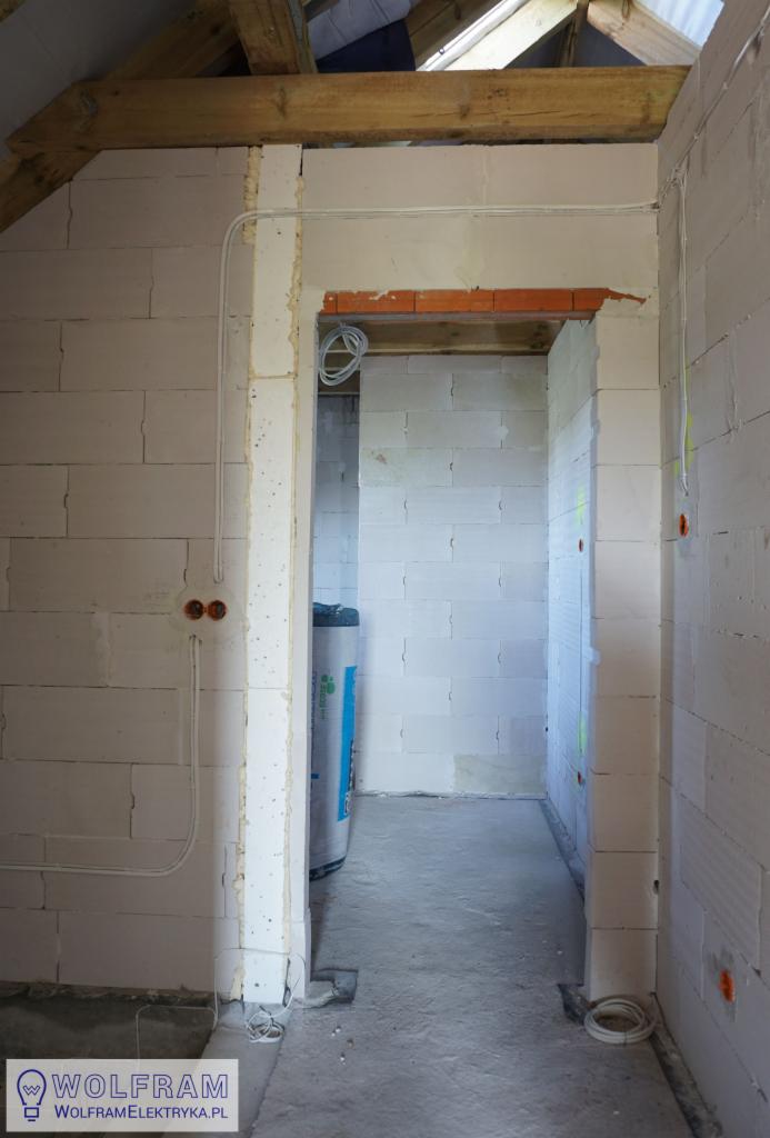 Wnętrze w trakcie prac instalacyjnych w surowym stanie: widoczne kable elektryczne prowadzone po ścianach z betonu komórkowego, puszki pod gniazdka, drewniana konstrukcja dachu w tle, oraz fragment...