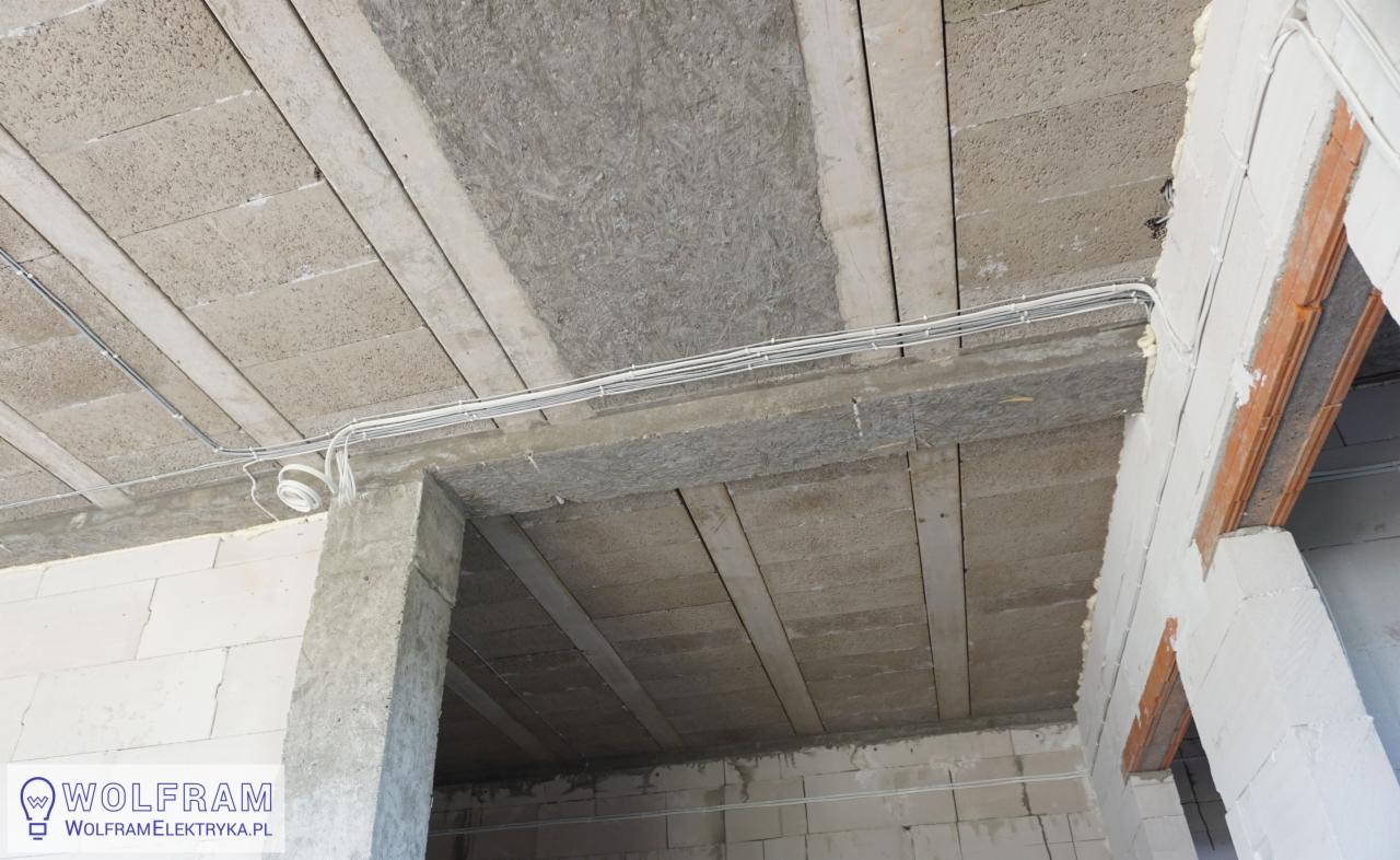 Instalacja elektryczna natynkowa w surowym wnętrzu: białe przewody ułożone na betonowym suficie z widoczną izolacją i elementami konstrukcyjnymi z betonu komórkowego.