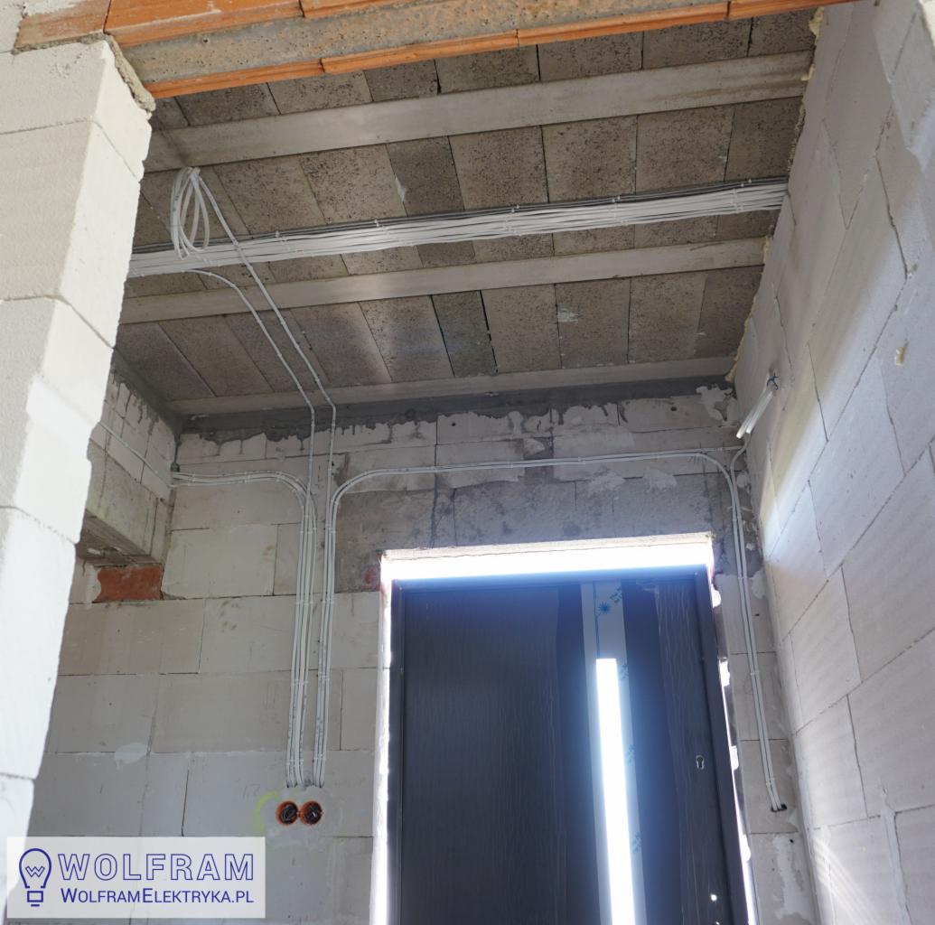 Surowe wnętrze w trakcie budowy z widocznymi przewodami elektrycznymi poprowadzonymi po ścianach z betonu komórkowego i pod stropem, prowadzącymi do puszki instalacyjnej. Widoczny fragment drzwi.