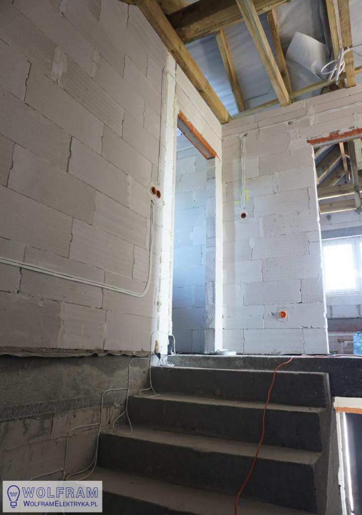 Wnętrze w trakcie prac, widoczne betonowe schody, ściany z białych bloczków, rozprowadzone kable elektryczne w peszlach oraz otwory pod gniazdka, drewniana konstrukcja dachu.