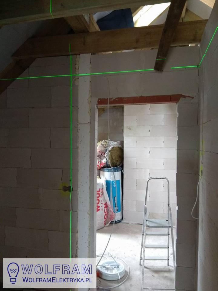 Wnętrze w trakcie prac wykończeniowych z widocznymi przewodami elektrycznymi, poziomica laserowa wyznacza pion i poziom na ścianach z betonu komórkowego, w tle widoczne materiały izolacyjne i drabina.