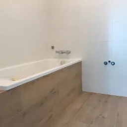 Nowa łazienka z wanną obudowaną panelami imitującymi drewno, białe płytki na ścianach, podłoga wyłożona panelami imitującymi drewno, widoczne przyłącza wodne i przewód elektryczny.