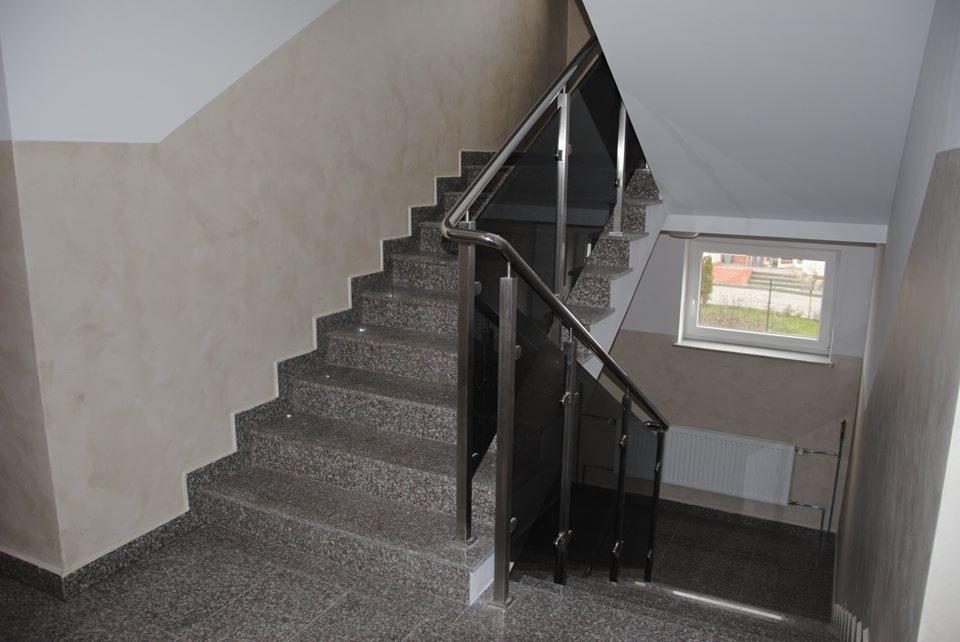 Klatka schodowa wykończona szarym, nakrapianym granitem, z balustradą ze stali nierdzewnej i czarnego szkła, oświetlona światłem dziennym wpadającym przez okno na półpiętrze.