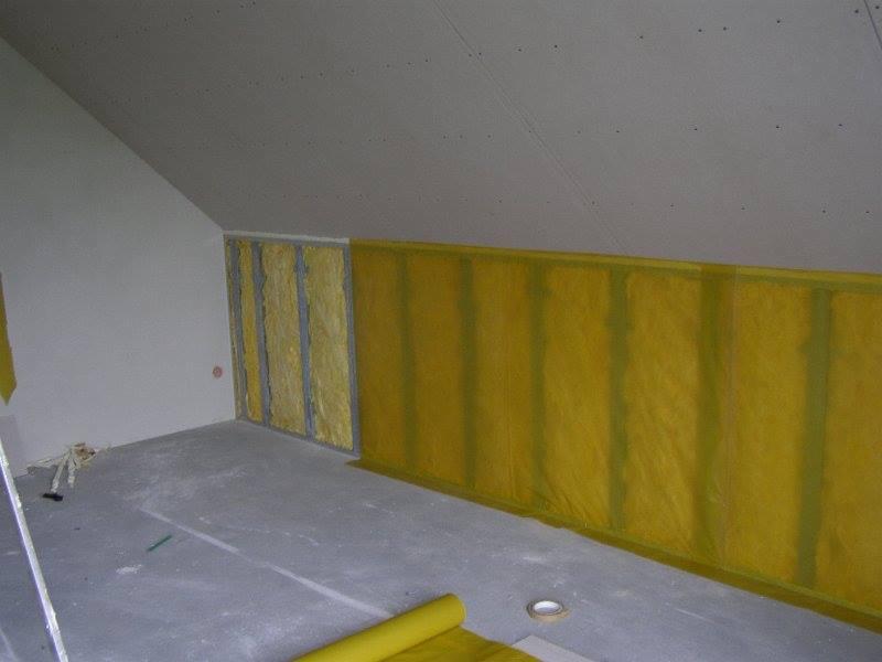 Poddasze w trakcie prac izolacyjnych: widoczne stelaże, wełna mineralna zabezpieczona żółtą folią oraz częściowo zamontowana płyta karton-gips na skosie sufitu.