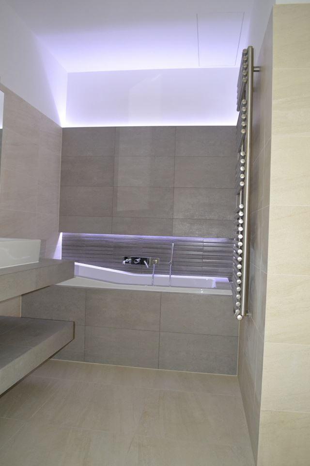 Nowoczesna łazienka z wanną obudowaną szarymi płytkami, podświetlona listwą LED, z grzejnikiem drabinkowym na ścianie i jasną podłogą.