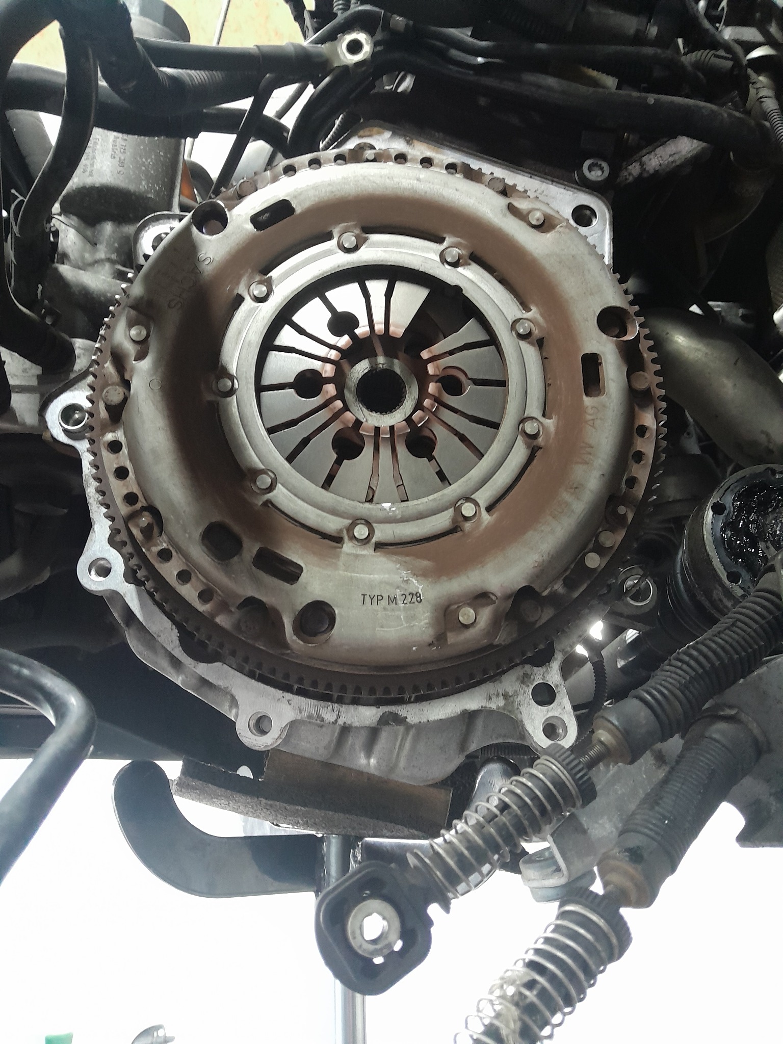 Sprzęgło samochodowe z widocznymi tarczami ciernymi, zębatym obwodem i oznaczeniem 'TYP M 228', zamontowane w silniku podczas naprawy.