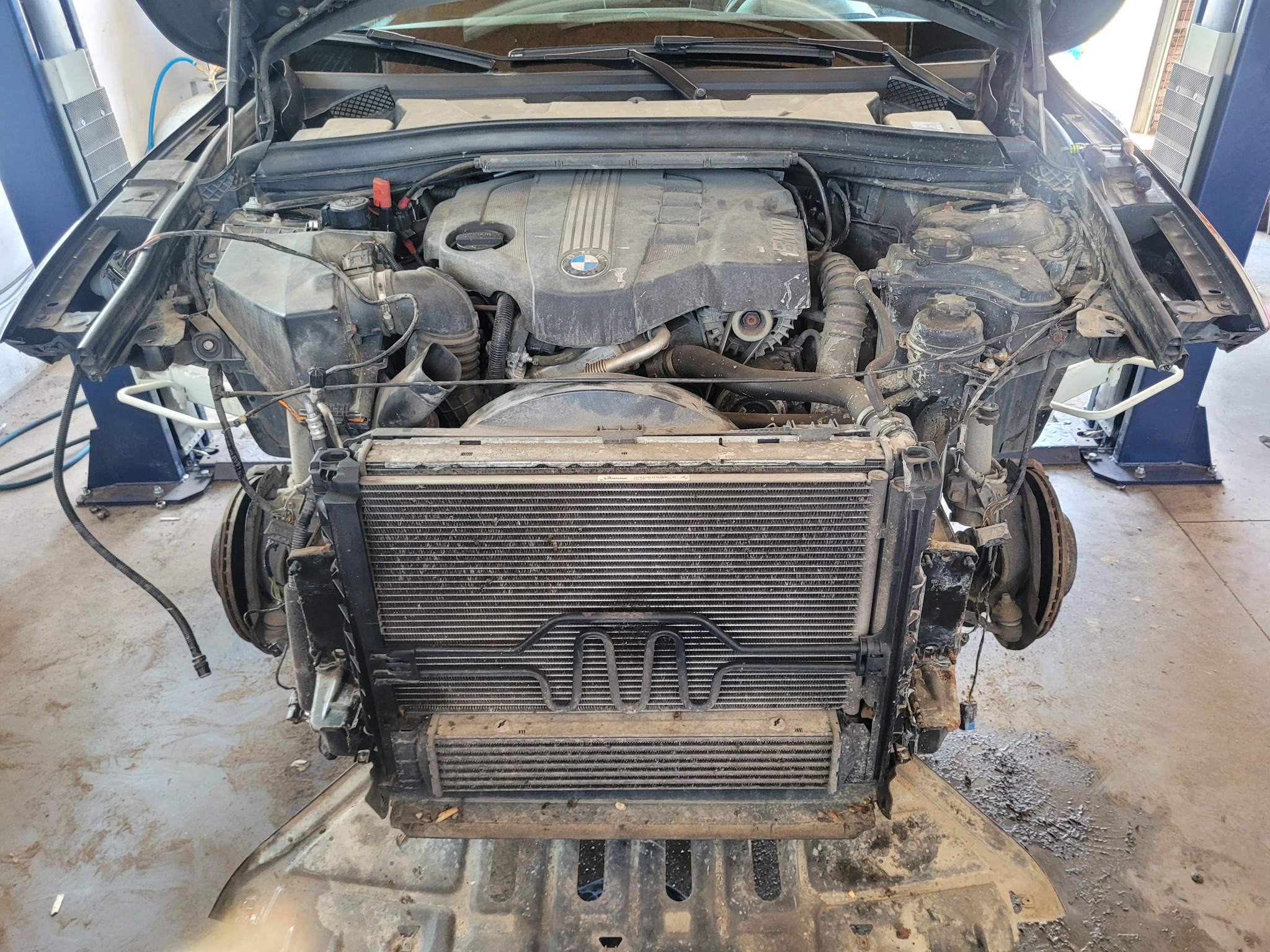 Uszkodzony przód samochodu BMW z widocznym silnikiem, chłodnicą i elementami zawieszenia w warsztacie.