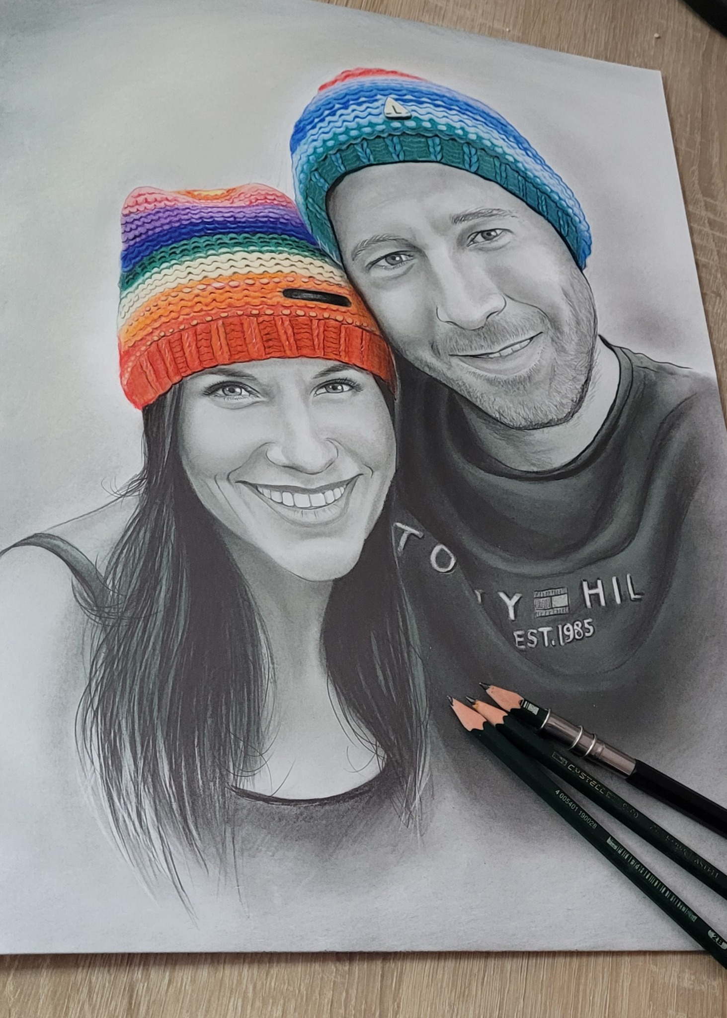 Ręcznie rysowany portret pary w czapkach, z widocznymi ołówkami na rysunku, tworzący osobisty i unikalny prezent. Detaliczna praca ołówkiem.