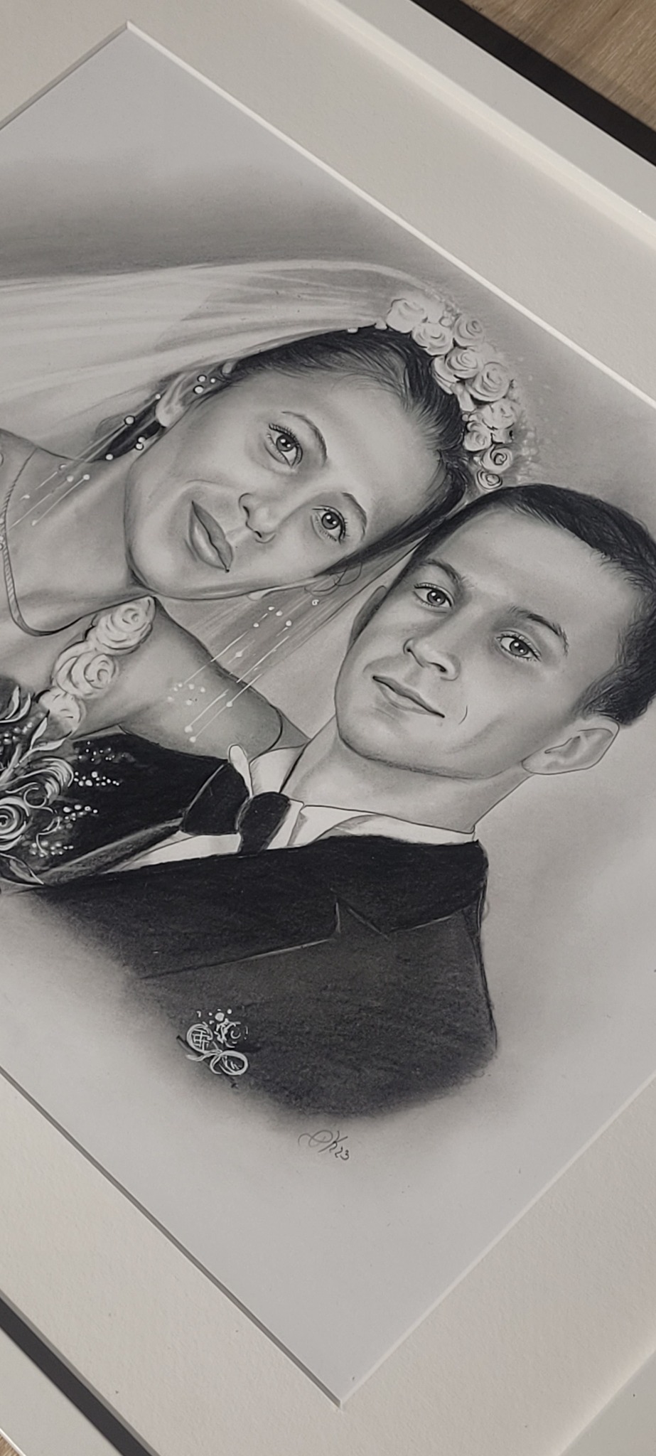 Ręcznie rysowany, oprawiony portret ślubny pary w czerni i bieli, z subtelnymi detalami i podpisem artysty w prawym dolnym rogu.