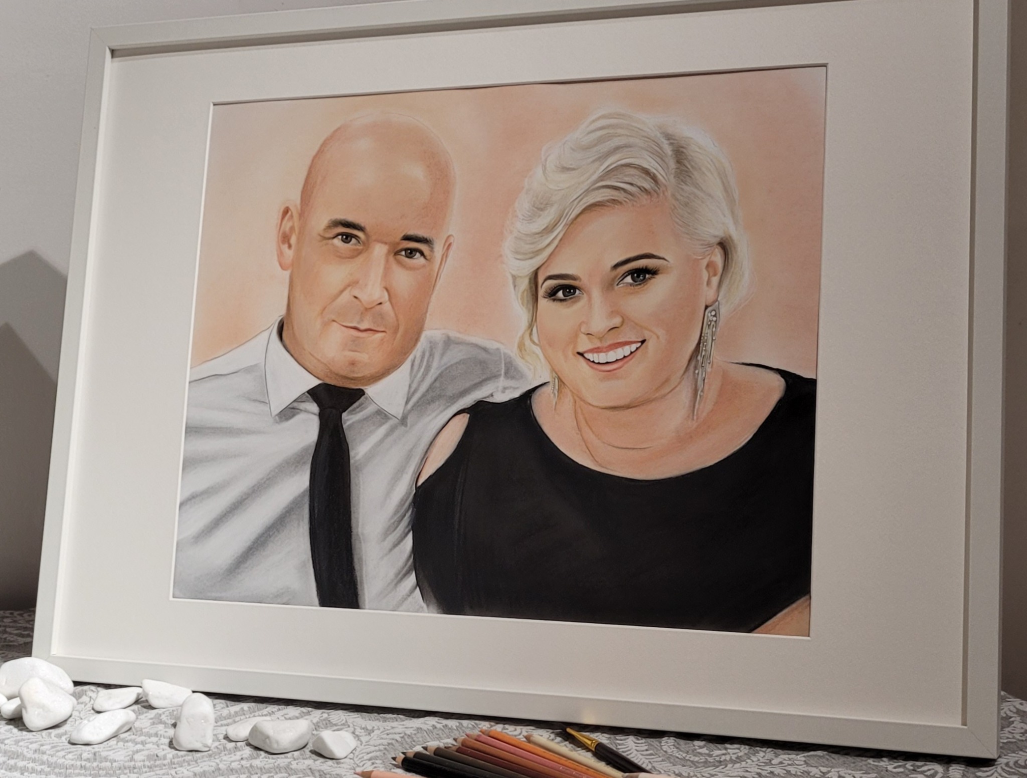Oprawiony portret pary, wykonany pastelami, leży na stole z kamieniami i kredkami. Ujęcie z boku podkreśla detale i fakturę.