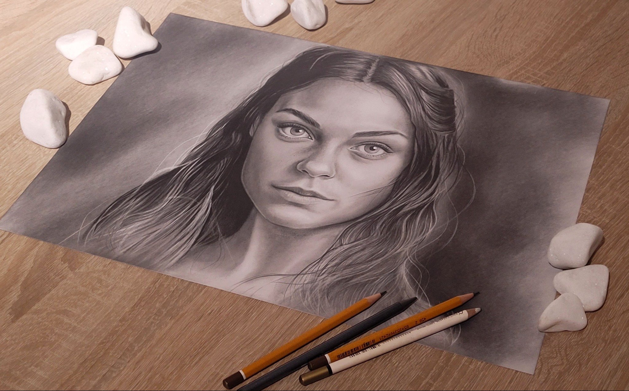 Portret kobiety, wykonany w technice rysunku ołówkiem, leży na drewnianym stole w otoczeniu kamieni i kredek. Realistyczne odwzorowanie detali.