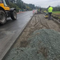 Prace drogowe: przygotowanie podłoża pod chodnik z użyciem ubijarki, w tle żółty traktor i pracownicy w odblaskowych kamizelkach.