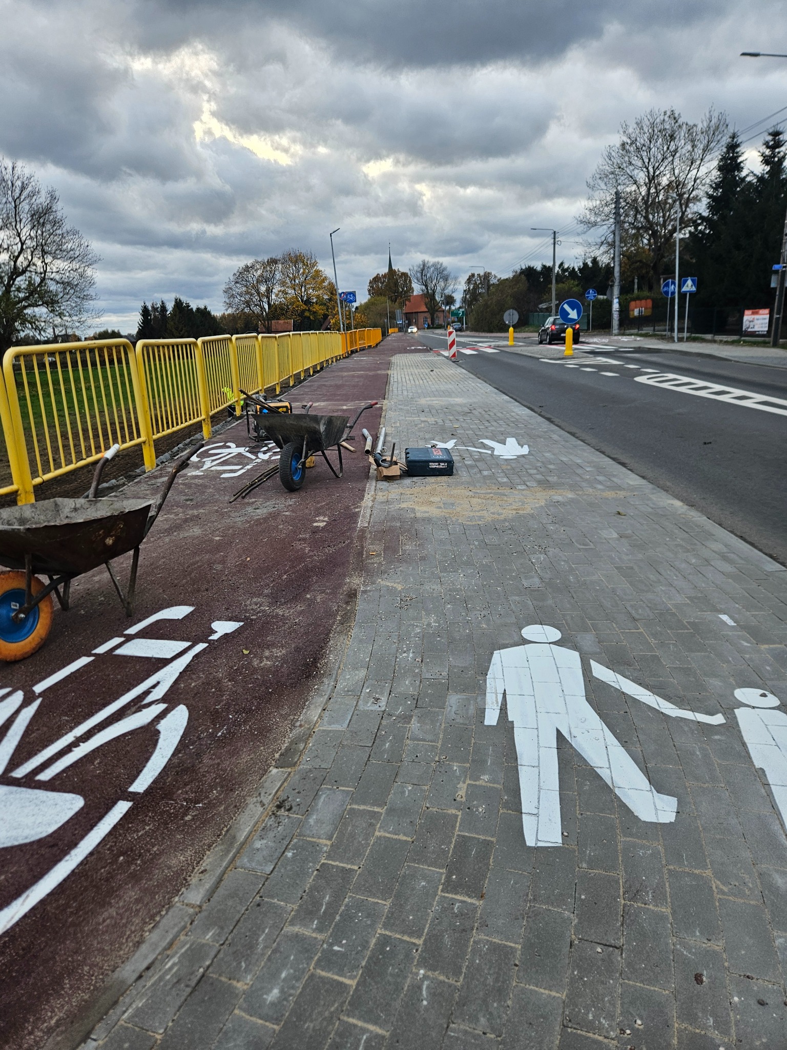 Nowo ułożona kostka brukowa z wymalowanymi białymi symbolami pieszych i rowerzystów, widoczne taczki i narzędzia na krawędzi ścieżki rowerowej, żółte barierki w tle.