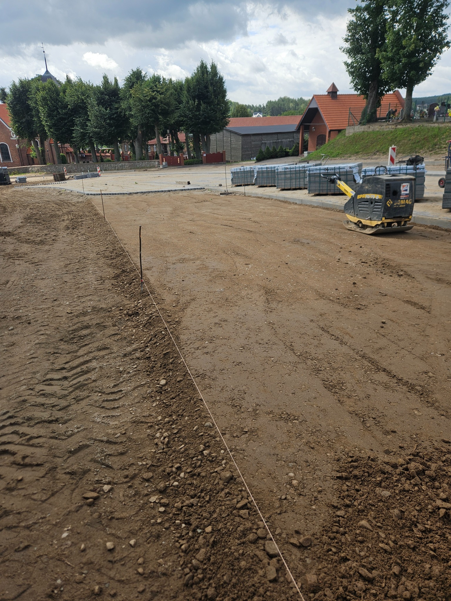 Przygotowany teren pod budowę z wytyczonymi granicami, ubijarką wibracyjną i paletami kostki brukowej w tle, widok na budynki w oddali.