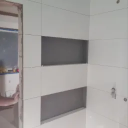 Ściana łazienki wyłożona białymi płytkami z wnękami pomalowanymi na szaro, widoczny fragment dłoni przy framudze drzwi.