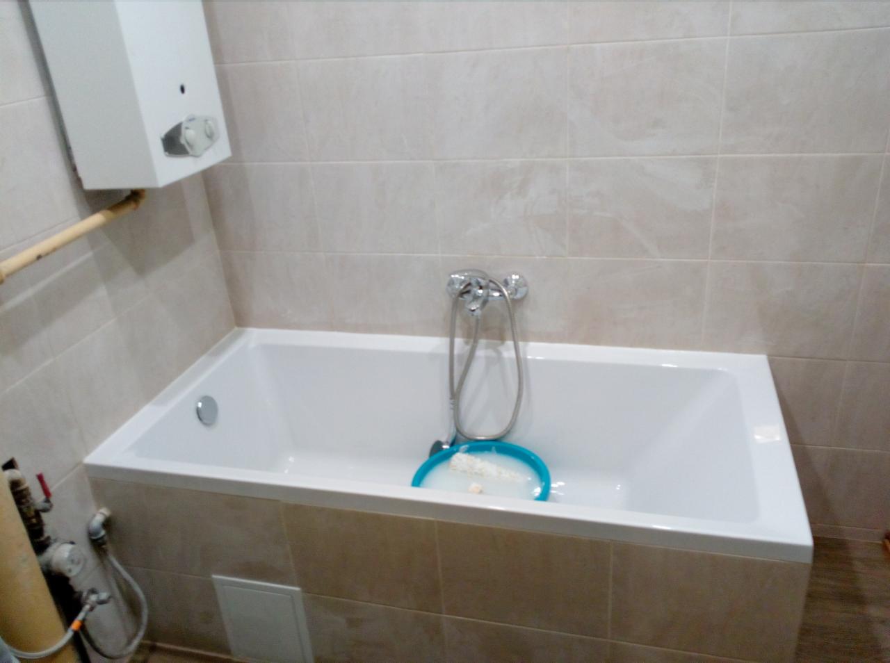 Biała wanna obudowana płytkami w łazience, chromowana bateria i słuchawka prysznicowa na ścianie, na wannie niebieska miska z gąbką, na ścianach jasne płytki.