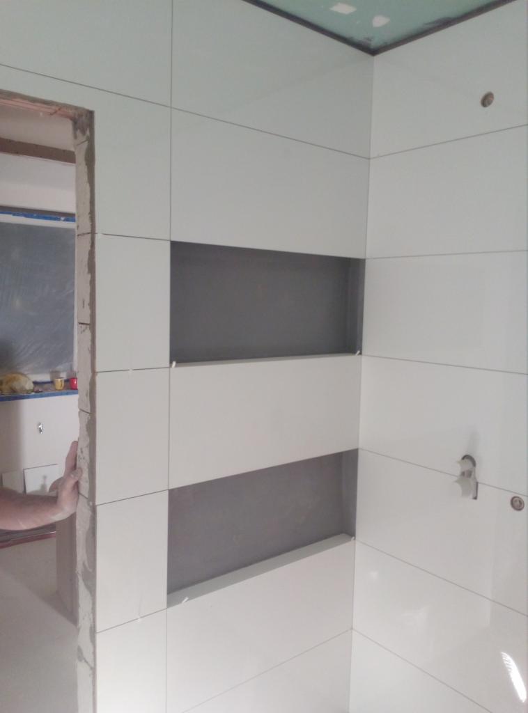 Ściana łazienki wyłożona białymi płytkami z wnękami pomalowanymi na szaro, widoczny fragment dłoni przy framudze drzwi.