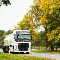 Cysterna firmy Petrodom przewożąca substancje niebezpieczne, widoczny pomarańczowy kod ostrzegawczy 33/1203 na białej tablicy, jadąca po asfaltowej drodze w otoczeniu drzew o jesiennych barwach.