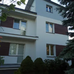 Nowoczesny dom z elewacją w kolorze białym i szarym, z drewnopodobnymi panelami, widoczne okna z odbiciami, balkon i iglaki w ogrodzie.