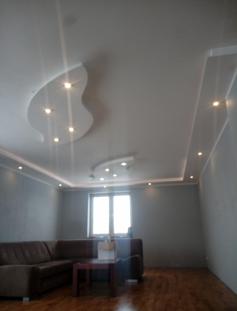 Nowoczesny salon z podwieszanym sufitem oświetlonym punktowymi lampami i listwami LED, szarymi ścianami i brązową sofą.