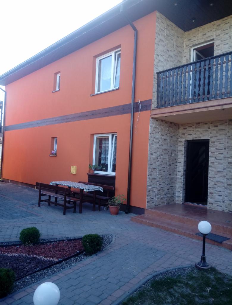 Dwukondygnacyjny dom z pomarańczową elewacją i fragmentem wykończonym kamieniem dekoracyjnym, widoczny balkon z czarną balustradą, na pierwszym planie kostka brukowa i małe krzewy.