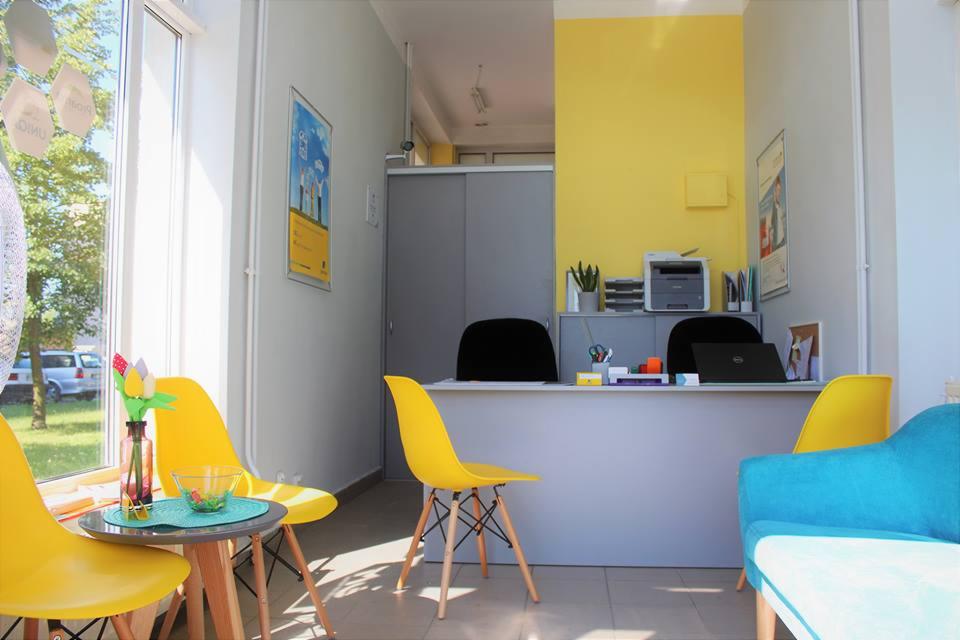 Jasne, nowoczesne biuro z żółtymi krzesłami i akcentem turkusowym, biurko z laptopem i drukarką, dekoracje okienne, kwiaty w wazonie na stoliku.